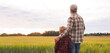 © Acronym - Farmer and his son in front of a sunset agricultural landscape. Man and a boy in a countryside field. Fatherhood, country life, farming and country lifestyle concept.