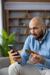 © Liubomir - Man reacting with a shocked expression, holding a credit card and smartphone, experiencing financial stress from an unexpected banking error or online fraud while sitting at home