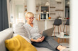 © Dorde - Senior woman in casual striped shirt and gray pants working on laptop in bright cozy living room