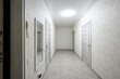 © evgeniykleymenov - Narrow hallway with light-colored textured wallpaper, grey tiled floor, three white doors, wall-mounted mirror and intercom, recessed ceiling light, minimalist design.