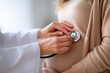 © maria_lh - Close-up of a doctor examining a pregnant woman's belly with a stethoscope. Prenatal medical check-up at the clinic. Maternity and healthcare concept