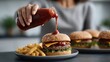 © alsu0112 - Hand pouring ketchup onto a cheeseburger with lettuce and cheese, accompanied by a side of golden French fries on a dark table in a modern kitchen setting