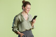 © Alliance - A woman in her 30s smiles down at her black smartphone. Dressed in muted green and gray, she stands against a light green backdrop, creating a professional, yet approachable feel.