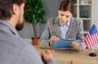 © Studio Romantic - Visa interview at US embassy office with officer reviewing applicant documents, writing notes with pen. Paperwork and immigration history review by lawyer sitting at desk with United States flag