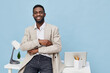 © SHOTPRIME STUDIO - Portrait of a smiling young African American man in casual blazer sitting at modern desk with laptop and stationery, looking confidently at camera. Studio shot with bright pastel background. People