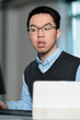 © fotolite - A young Asian works at his laptop in a contemporary office setting, ideal for workplace productivity, professional development, entry level employment, or corporate work environment concepts.