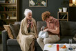 © AnnaStills - Senior Caucasian woman sitting on sofa holding mug while young Caucasian teenager sitting beside her searching inside bag, medicine bottles and fruit on table in foreground