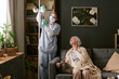 © AnnaStills - Senior Caucasian woman sitting on sofa looking up at young adult Caucasian female nurse adjusting IV stand in living room setting, medical care at home concept