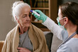 © AnnaStills - Senior Caucasian woman sitting while female healthcare worker measuring forehead temperature with digital thermometer, medical examination taking place in clinical setting