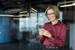 © Liubomir - Young businesswoman standing in a contemporary office, smiling while typing on her smartphone, engaging with mobile technology and digital communication