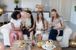 © wavebreak3 - Diverse moms group sitting on couch holding baby sharing smartphone near cake, balloons