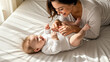 © Celt Studio - Happy mother playing with laughing baby lying on white bed in sunny bedroom enjoying family time and motherhood love together