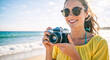 © NeuroCake - Happy young woman with a vintage camera on a sunny beach. Smiling tourist enjoying summer vacation and her photography hobby by the ocean. Travel and leisure lifestyle concept