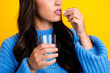 © deagreez - Young mixed race woman in blue sweater taking pill with glass of water against yellow background showcasing casual lifestyle and fashion trend