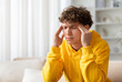 © Anastasiya - Teenage boy touching head with closed eyes, sitting on couch at home, wearing yellow hoodie, experiencing headache, stress, or mental fatigue in everyday lifestyle indoors