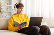 © Anastasiya - Smiling teenage guy wearing yellow hoodie and headphones, studying at home with laptop and notebook, writing notes, enjoying online learning, focus, and comfortable modern lifestyle indoors