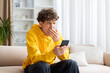 © Anastasiya - Tired teenage guy yawning while looking at smartphone, sitting on sofa at home in yellow hoodie, showing fatigue, boredom, and everyday digital routine indoors