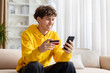 © Anastasiya - Teenage guy smiling while checking smartphone and holding credit card, sitting on sofa at home, wearing yellow hoodie, enjoying online shopping and digital lifestyle indoors