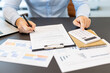 © Liubomir - Business person analyzing financial data, calculating numbers using a calculator, and writing on a document with various charts and graphs on the desk, focusing on accounting and tax preparation