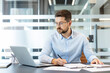 © Liubomir - Young businessman wearing glasses sitting at desk in a contemporary office, carefully focusing on calculations and paperwork while using a laptop to manage data and financial reports