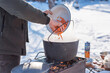 © T.Den_Team - Person preparing hot food outdoors during winter using a metal cauldron over open fire. Close view of hands cooking a meal in cold weather