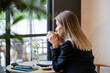 © DimaBerlin - Pensive businesswoman freelancer enjoying aromatic coffee in stylish restaurant on lunch break during workday. Confident female drinking from cup looking outside comfortable sitting at table in cafe.