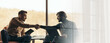© Prostock-studio - Two men sit in a modern workspace. One man shakes hands with the other while sitting by a window. Sunlight enters the room, creating a relaxed atmosphere for discussion and agreement.