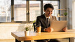 © SHOTPRIME STUDIO - Young businessman in suit smiling while using laptop at wooden desk with coffee cup and plant near large windows in bright modern office. Concept for remote work and productivity.