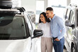 © Prostock-studio - Happy middle-eastern young family choosing new auto in salon, standing by nice white vehicle, looking inside car through window, having conversation, embracing and smiling, copy space