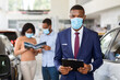 © Prostock-studio - Portrait Of Handsome Black Car Salesman Wearing Protective Medical Mask At Workplace In Showroom, Young Dealership Center Manager With Clipboard In Hands Helping Couple To Purchase Family Vehicle