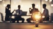 © Ikhwan - Illuminated Light Bulb on Table with Blurred Business People in Background.