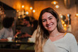 © Jose Calsina - A joyful young woman smiles brightly while enjoying her time at a trendy cafe, radiating happiness and warmth as she interacts with her friends in the inviting atmosphere of the bar.