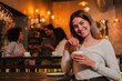 © Jose Calsina - A cheerful young woman savoring a delicious cup of coffee while smiling and interacting with friends in a lively cafe atmosphere, radiating joy and warmth in her delightful experience.
