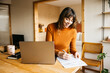 © Jordi Salas - Woman smiling while working at home, using a laptop and writing on documents