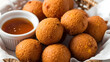 © Hammad - A basket of golden-brown, crispy fried hushpuppies served with a small bowl of honey or syrup for dipping, presented in a rustic setting.