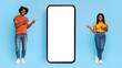 © Prostock-studio - Happy black millennial couple in casual standing by huge brand new cellphone with blank screen and pointing at gadget, recommending newest mobile app, mockup, blue studio background, panorama