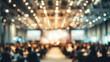 © Subin - Blurry background of a busy conference hall filled with attendees and illuminated by bright overhead lights.