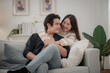 © surachetkhamsuk - Happy young Asian couple hugging, sitting on cozy couch together, overjoyed laughing woman and man having fun, enjoying leisure time, relaxing on sofa in living room at home