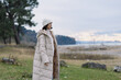 © SHOTPRIME STUDIO - Woman in a long beige puffer coat and hat standing near a lake, autumn park landscape, calm sky, distant trees, soft light, outdoor fashion and travel mood.