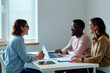 © Mediaphotos - Young woman interviewing Black young man and young woman sitting at table using laptop and tablet smiling and engaging in conversation