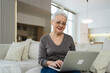 © Юлия Завалишина - Confident stylish happy middle aged senior woman using laptop at home. Stylish older mature 60s grandmother sitting at couch looking at computer screen typing chatting reading writing email