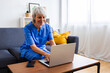 © Xavier Lorenzo - Portrait of senior healthcare worker in blue scrubs working on laptop sitting on sofa at home. Technology and healthcare business concept.