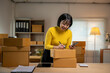 © Wasana - Young woman managing sme business writing shipping information on a box and using her cell phone