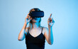 © Volodymyr_sh - Young woman using VR headset and pointing in virtual space, immersive gaming and interactive technology experience on blue background