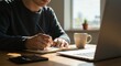 © Cleverson - A man sitting at a desk writing in a notebook with a laptop and coffee cup nearby