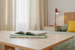 © AnnaStills - Stack of books with open notebook lying on wooden desk in front of bed with green bedding and yellow pillow, sunlight streaming through window with curtains in background