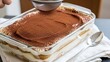 © Skylover - A hand sifting cocoa powder onto a homemade tiramisu dessert in a clear glass baking dish with a spoon on a white napkin