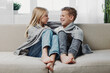 © SHOTPRIME STUDIO - Two children wrapped in grey blankets sit barefoot on a sofa facing each other, smiling and enjoying a cozy moment in a bright living room with plants in the background.