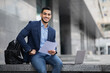 © Prostock-studio - Handsome arab entrepreneur sitting by office building with laptop, holding documents and smiling, copy space. Confident cheerful guy in suit getting ready before business meeting, reading speech