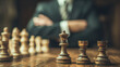 © Sergej Gerasimov - Strategic wooden chess pieces positioned on a table with a businessman in suit contemplating next move in blurred background for decision making concept